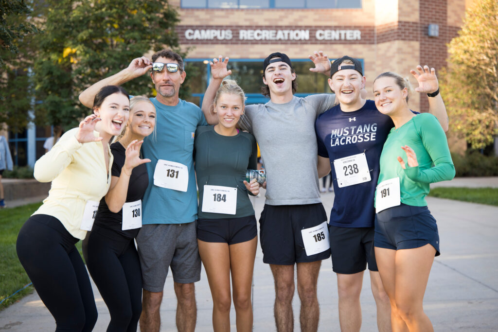 A group of six people wearing race numbers pose cheerfully outdoors in front of a campus recreation center building.
