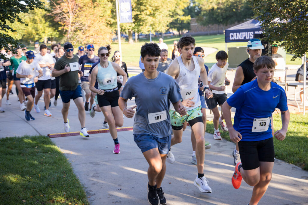 A group of runners participates in an outdoor race on a paved path lined with trees and grass.