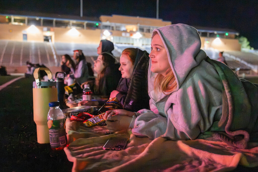 A group of people sit on blankets at a nighttime outdoor event, enjoying snacks and drinks while watching something together.