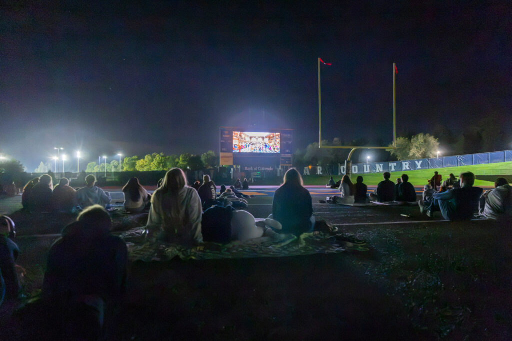 A group of people sit on blankets at night on a sports field, watching a large screen displaying a live event.