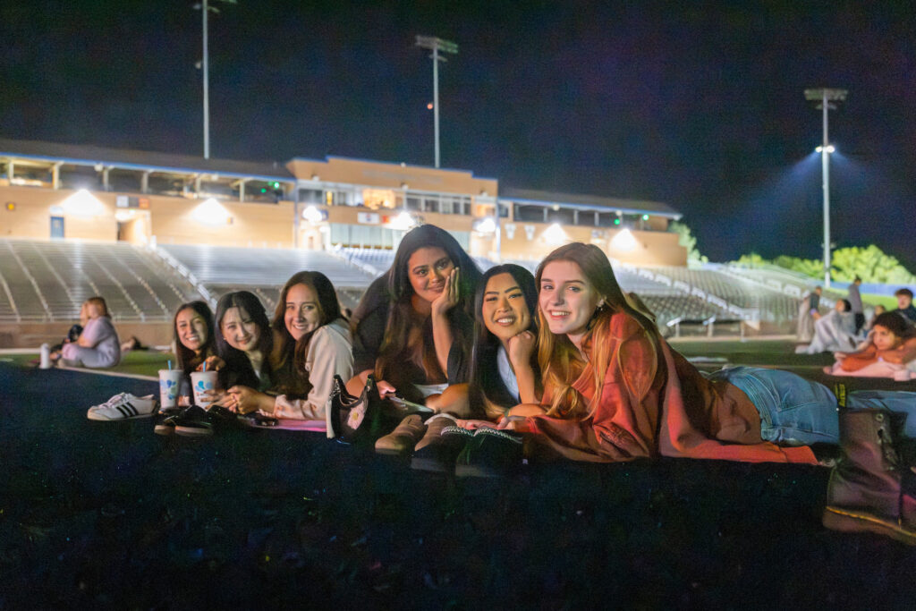 A group of young people relax on a blanket at a nighttime sports field, smiling for the camera under stadium lights.