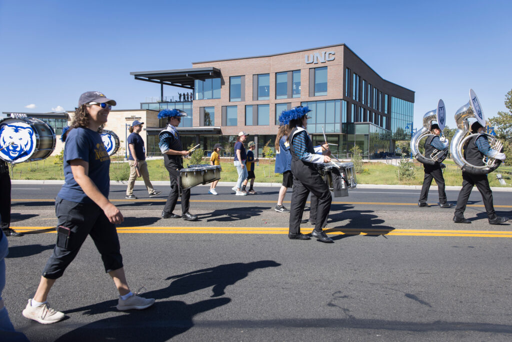 A marching band in blue and black uniforms with feathered headpieces performs on a street in front of a modern building marked "UNC" under a clear blue sky.