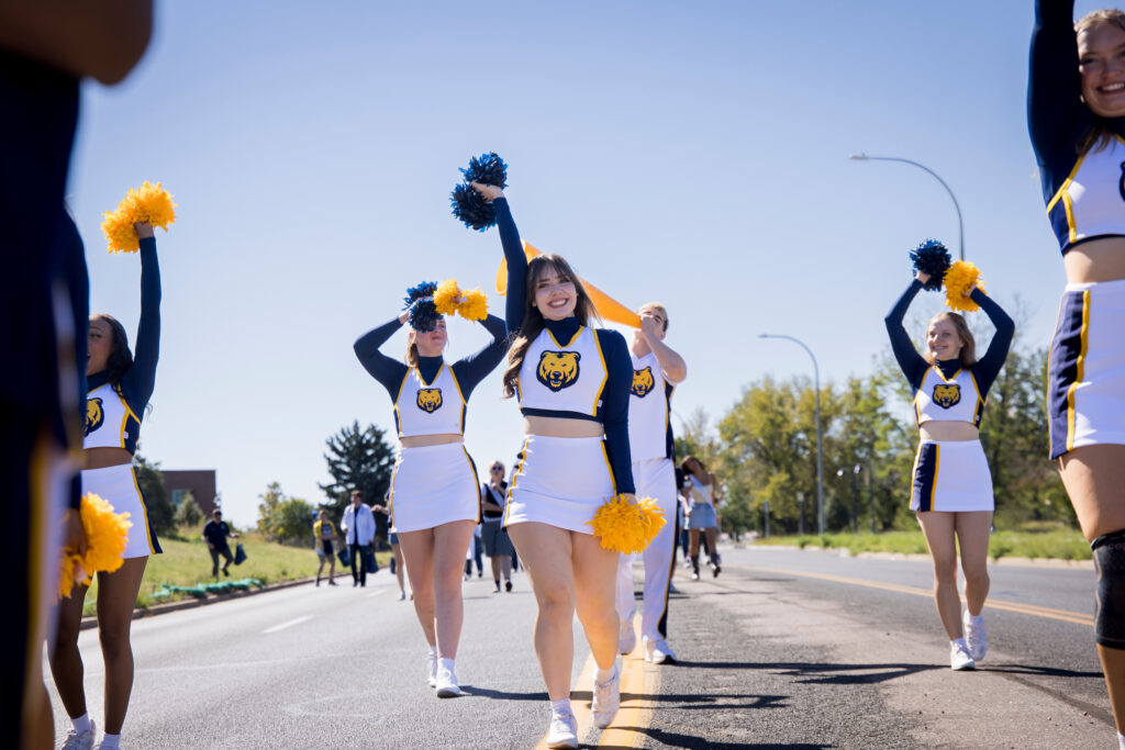 Cheerleaders in blue and yellow uniforms march down a sunny road, waving pom-poms and smiling.