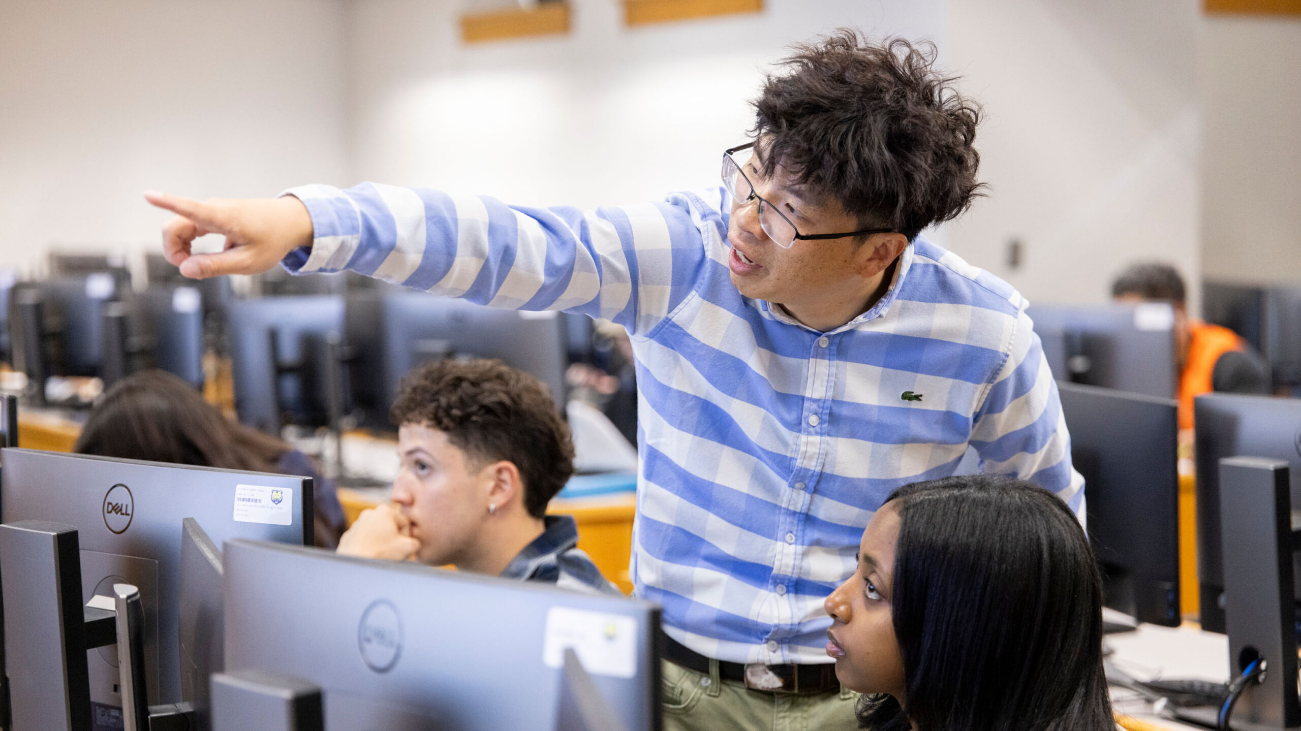 A teacher in a blue and white striped shirt is pointing and explaining something to two students sitting at computer desks. The students appear engaged and focused on the lesson. The classroom is equipped with multiple computer stations.