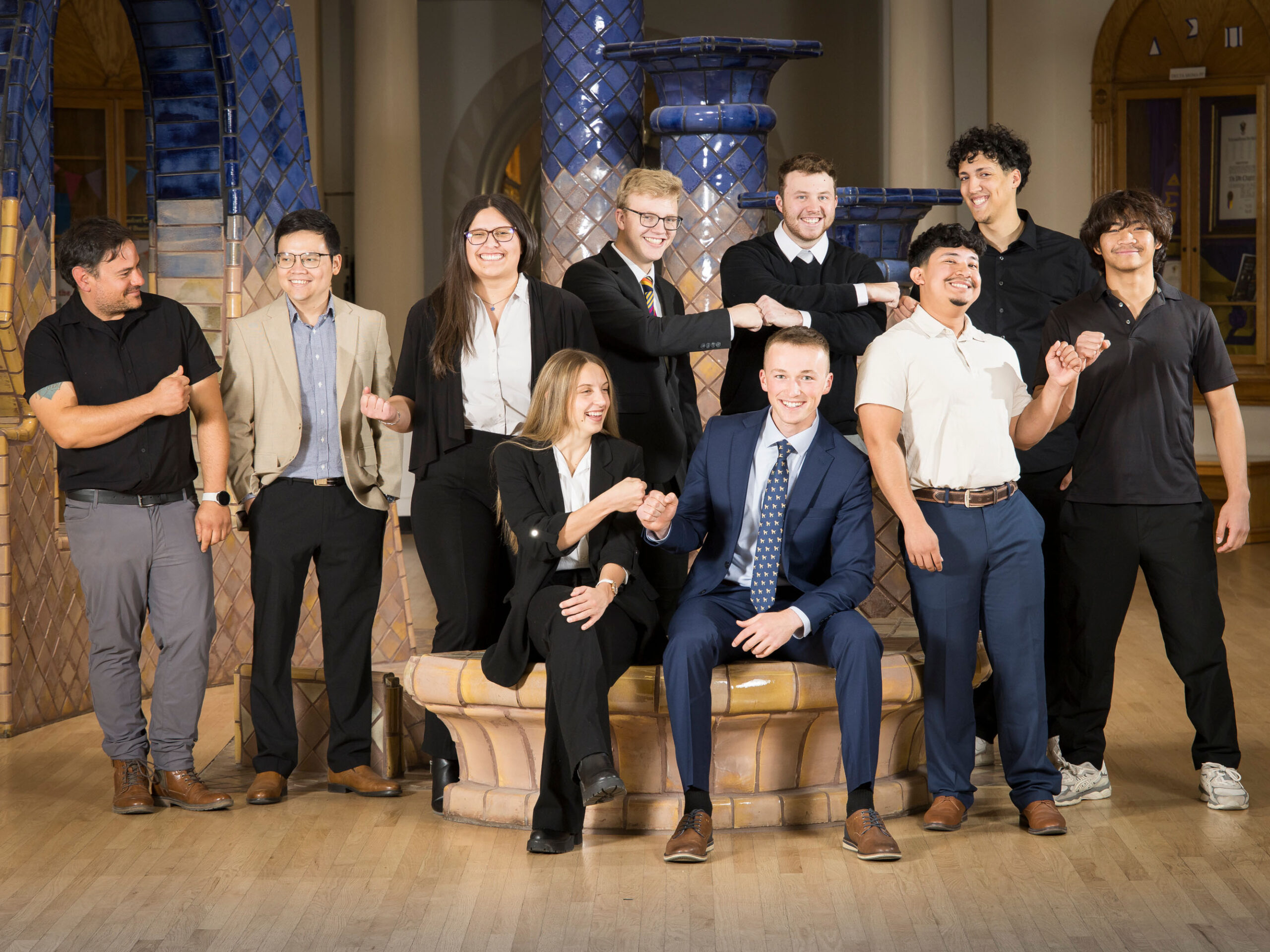 A group of ten people is gathered indoors, smiling and interacting with each other. They are dressed in a mix of formal and casual attire, with some sitting on a tiled bench and others standing around. The setting includes decorative blue pillars.