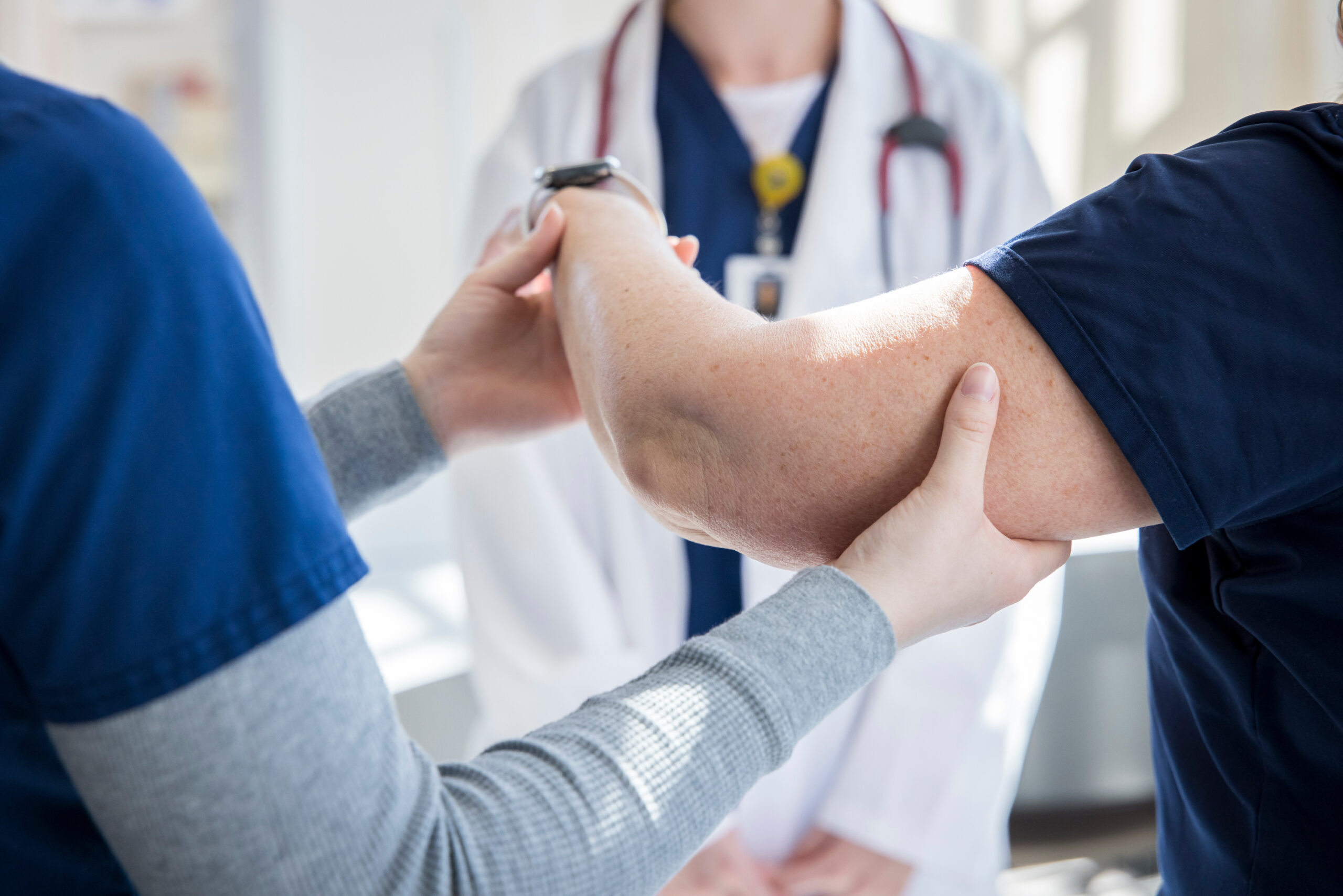 A student examines a patient's elbow with a supervising doctor in the background.