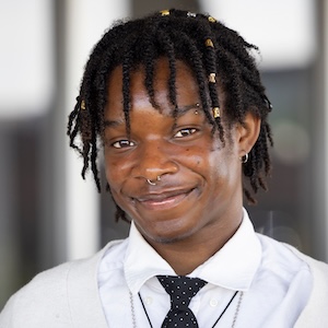A person with dreadlocks adorned with gold beads smiles while wearing a white shirt, black polka-dot tie, and a light-colored cardigan with a prominent circular pendant necklace.