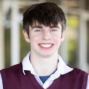 A young person with short dark hair and blue eyes smiles warmly while wearing a maroon sweater vest over a white collared shirt.