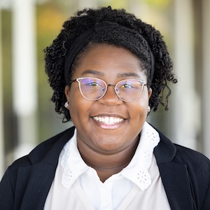 A smiling person with curly hair, glasses, and a black headband wears a white blouse under a black blazer.