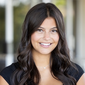 A smiling person with long dark wavy hair wearing a black top and a delicate necklace, standing outdoors with a blurred background.