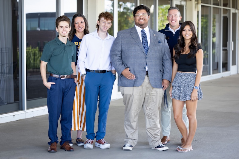 A group of six people pose together outdoors in front of a modern building with large glass windows.