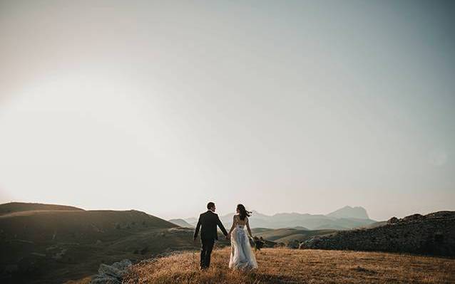 A heterosexual couple dressed in wedding clothing and facing a mountain range holds hands.