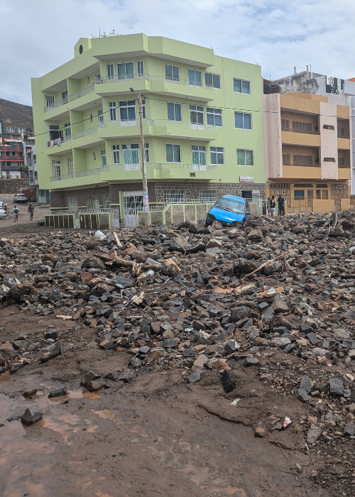 A green building with rubble, debris, and a wrecked car in front of it