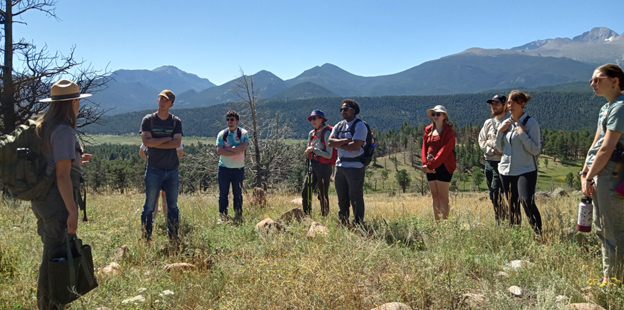 Eight students standing in front of a mountain range talking to a park ranger.