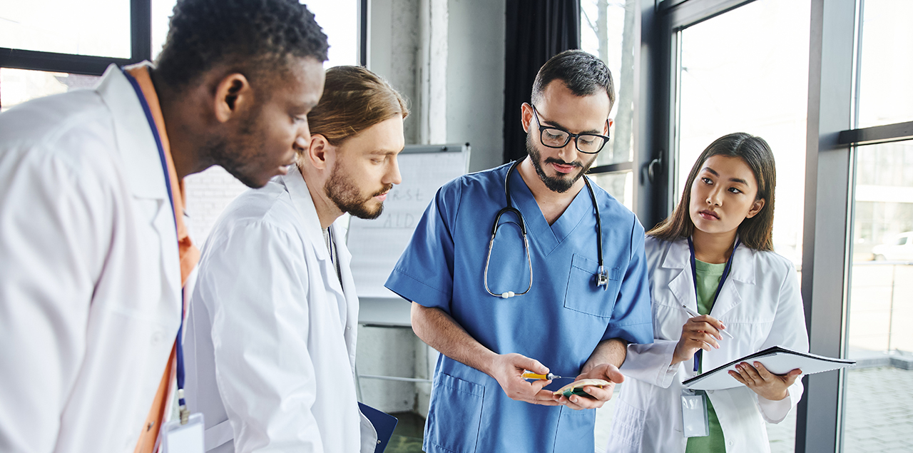 young male and female doctors in white lab coats taking notes and learning from older doctor in blue scrubs.