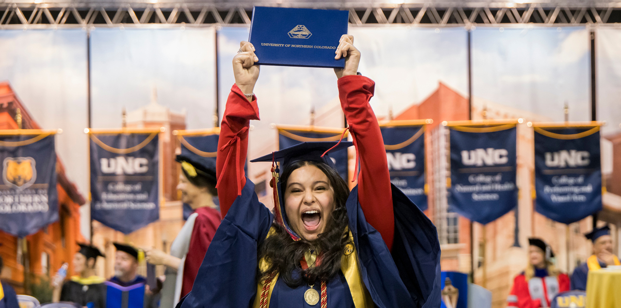 Student in cap and gown at graduation holding degree above her head and celebrating