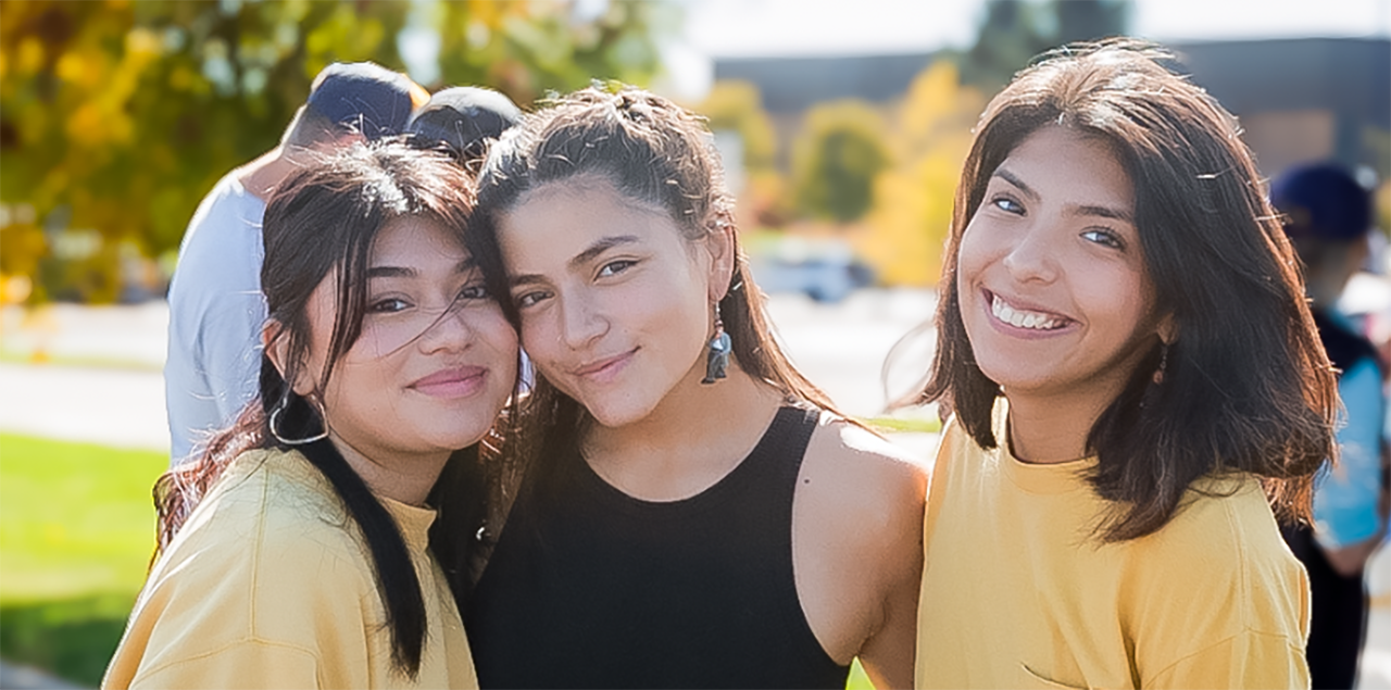 Tres sonrientes estudiantes universitarias permanecen juntas y de pie en el exterior durante un acto para celebrar las comunidades hispanas/latinas. 