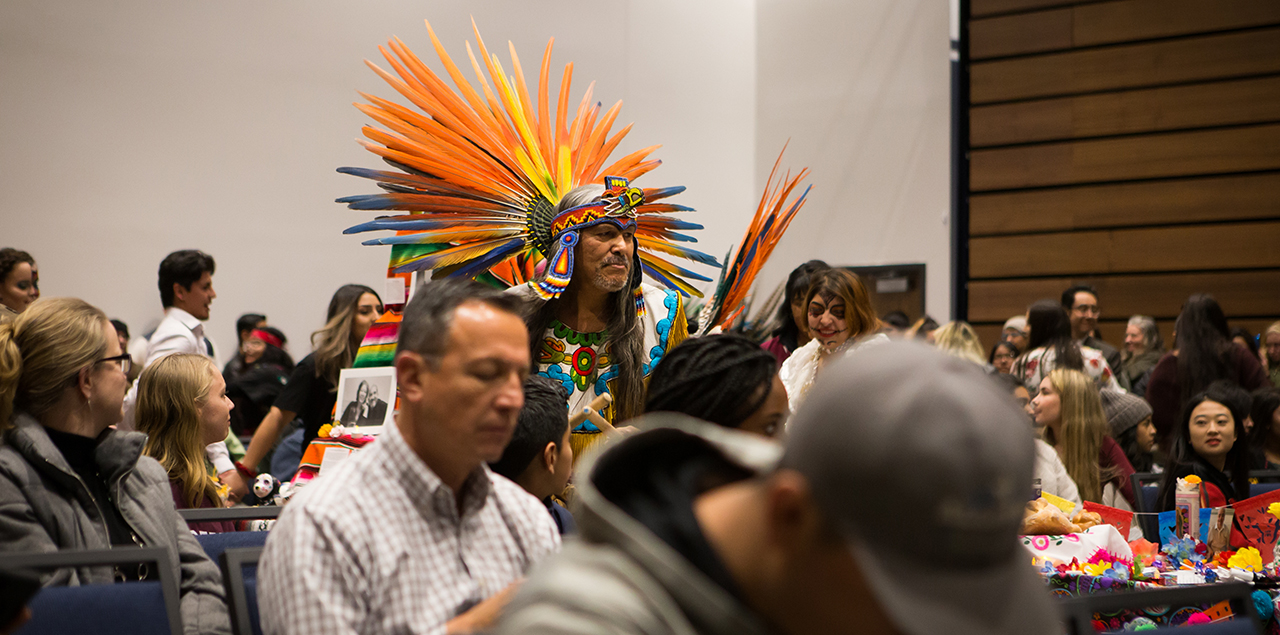 Photo of people sitting at tables and a man wearing a penacho, an aztec feathered headdress