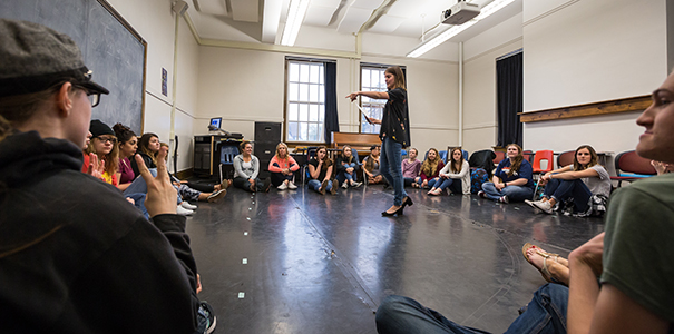 instructor standing in circle of students in a large room conducting acting class