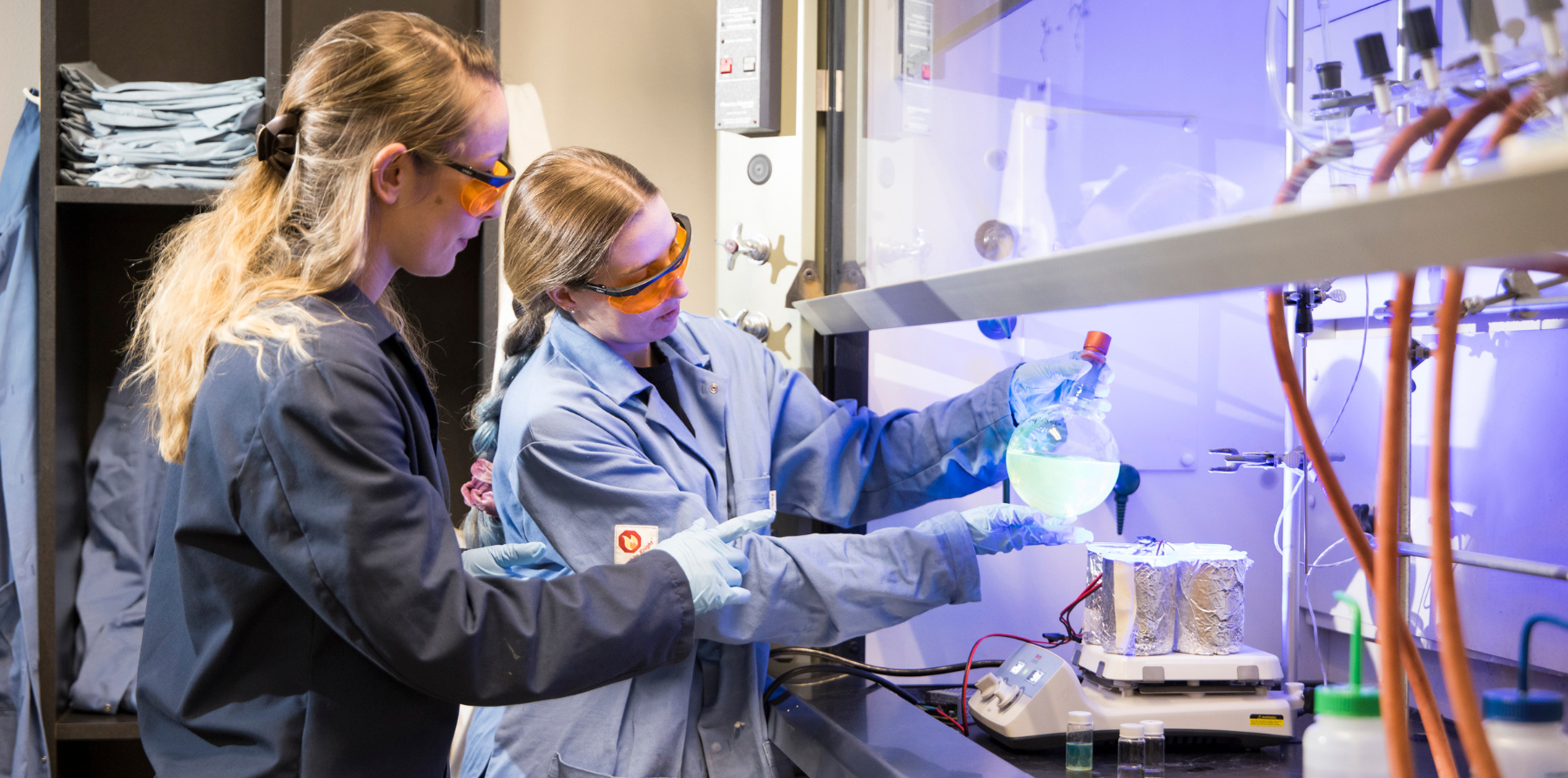A pair of women in lab coats and goggles working with chemicals.