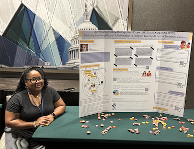 Kouston Worthington, a black woman in her 30s with long dark hair wearing a black shirt and black striped pants, sits beside a table with a poster presentation on it, along with some candy spread out on the table.