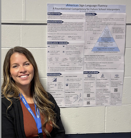 Director, Dr. Emily Girardin, a woman in her 30s with brown hair, a brown shirt, and a black sweater stands beside a poster at the RID conference