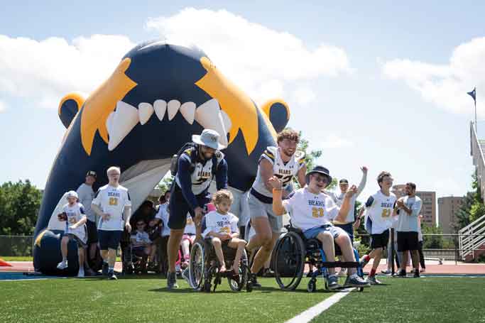 UNC football student-athletes running onto the field, accompanied by disabled children during the No Limits Camp, fostering inclusivity and joy.